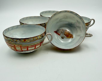 Könnte beinhalten: Satz von vier Vintage-Teetassen mit hellblauem Innenraum und dekorativem Rand. Jede Tasse hat ein einzigartiges Design, darunter ein rot-goldenes Brückenmotiv und eine Fächerillustration. Die Tassen haben silberne Griffe.