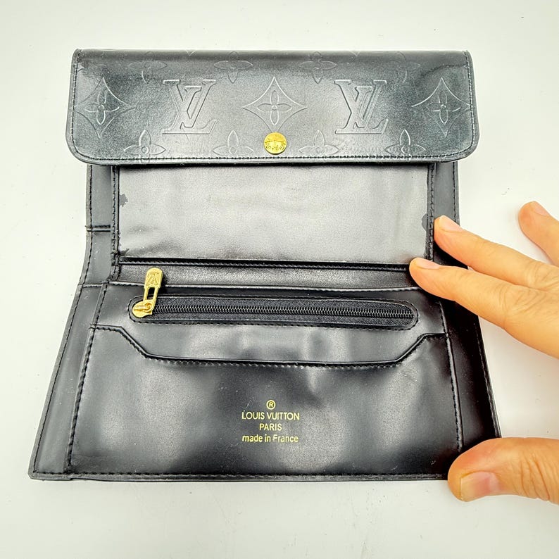 Puede incluir: Cartera de cuero negro con estampado de monograma en relieve y herrajes dorados. La cartera presenta un cierre de solapa con broche, un compartimento con cremallera y el texto "LOUIS VUITTON PARIS made in France".