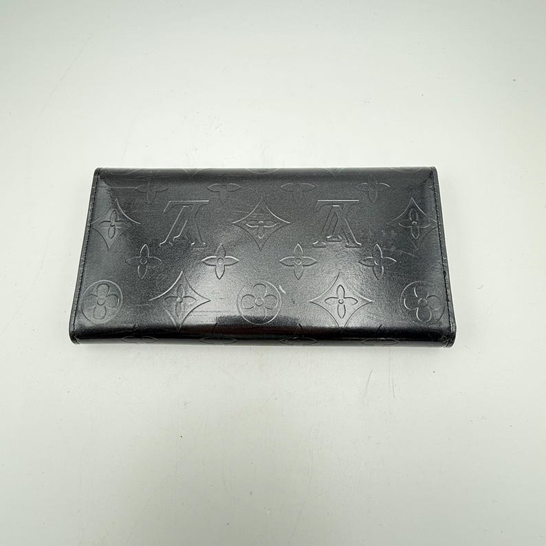 Puede incluir: Cartera de cuero negro con estampado de monograma en relieve. La cartera rectangular presenta un patr&oacute;n repetido del logotipo de la marca. El cuero tiene un acabado brillante y la cartera parece estar cerrada.