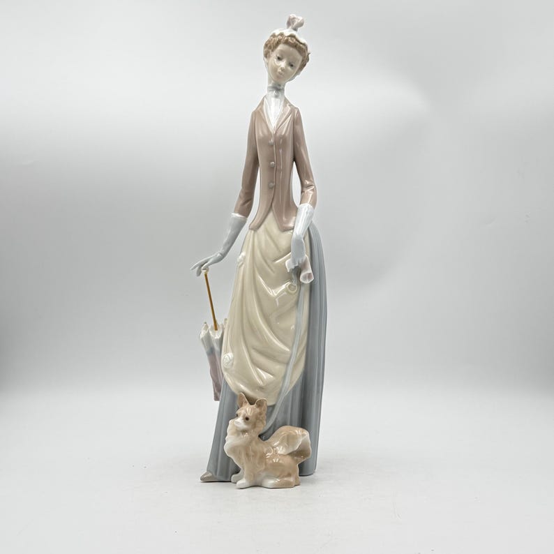 May include: A porcelain figurine of a woman in a long dress and jacket, holding an umbrella and a rolled paper. A small dog sits at her feet. The figurine is primarily in shades of beige, gray, and brown.