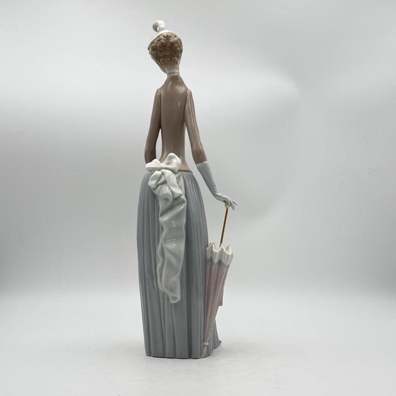 May include: A porcelain figurine of a woman in a long, pleated gray skirt and a light brown top. She wears white gloves and holds a pink and white umbrella. A large white bow adorns the back of her skirt. The figurine has a delicate, elegant design.