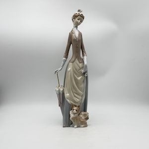 May include: A porcelain figurine of a woman in a long dress, holding an umbrella. The woman is wearing a brown jacket and a long, light-colored skirt. A small dog figurine is at her feet. The figurine is a decorative item.