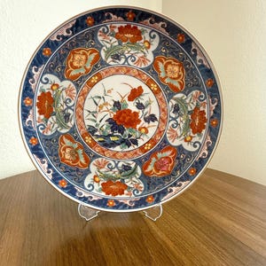 May include: A decorative ceramic plate with a floral design. The plate features a blue border with orange and white floral patterns. The center of the plate has a detailed floral arrangement with red and blue accents. The plate is displayed on a clear stand.