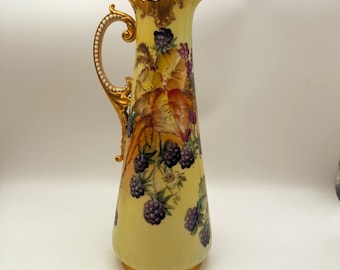 May include: A tall, yellow ceramic pitcher with a gold handle and rim. The pitcher is decorated with a painted design of blackberries and leaves. The handle is adorned with gold accents and small white beads.