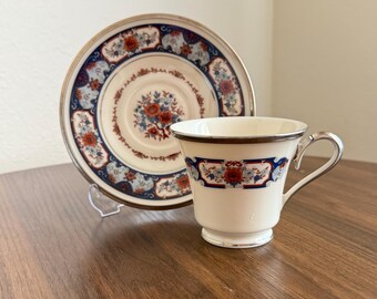 May include: A white teacup and saucer set with a floral design. The cup and saucer have a blue and red floral pattern with gold trim. The cup has a silver handle and a matching band of the floral design.