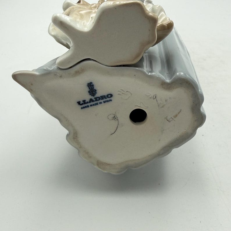 May include: A porcelain figurine with a light blue dress and a tan-colored headpiece. The bottom of the figurine is visible, displaying the Lladro logo and the text "Hand Made in Spain."