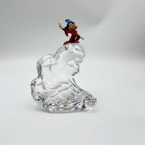 Op de afbeelding: Een heldere glazen sculptuur met een golfachtig ontwerp, bekroond met een klein beeldje van Mickey Mouse in een tovenaarsoutfit. Het beeldje draagt een blauwe hoed en een rode mantel. De sculptuur staat op een witte achtergrond.