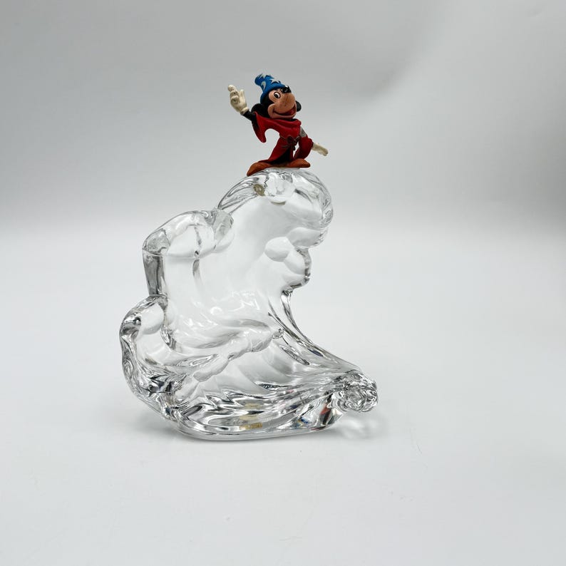 Op de afbeelding: Een heldere glazen sculptuur met een golfachtig ontwerp, bekroond met een klein beeldje van Mickey Mouse in een tovenaarsoutfit. Het beeldje draagt een blauwe hoed en een rode mantel. De sculptuur staat op een witte achtergrond.