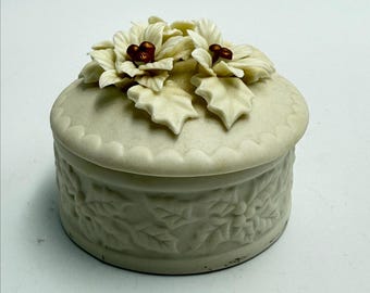 May include: A cream-colored, round trinket box with a lid. The lid is adorned with sculpted white flowers and brown accents. The box's body features a raised floral pattern. The box is a decorative item.
