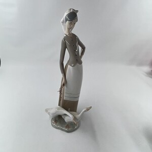 May include: A porcelain figurine depicting a woman in a long dress and head covering, holding a bundle. Two white geese rest at her feet on a gray base. The woman's outfit is gray and white.