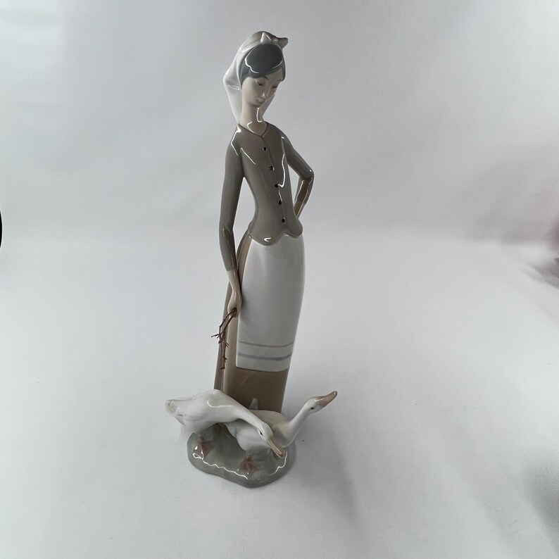 May include: A porcelain figurine depicting a woman in a long dress and head covering, holding a bundle. Two white geese rest at her feet on a gray base. The woman's outfit is gray and white.