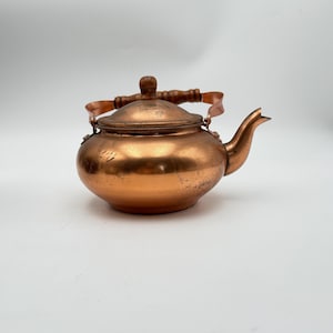 May include: A round copper teapot featuring a polished metallic finish with visible surface wear and scratches. The teapot includes a matching copper lid with a central knob and a curved spout, complemented by a dark brown wooden handle.