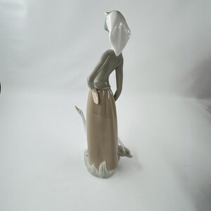 May include: A porcelain figurine of a woman in a long skirt, with a white head covering, and a goose. The woman is in a relaxed pose, with one arm resting on her hip. The figurine is primarily in shades of gray, beige, and white.