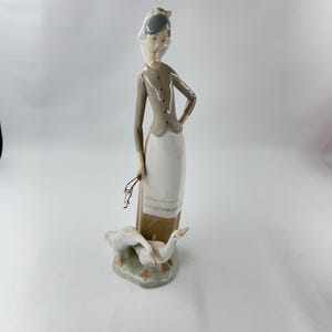 May include: A porcelain figurine of a woman in a long dress and head covering, holding a small branch. Two white geese are at her feet, on a small, green base. The woman's outfit is tan and white, with a gray jacket.