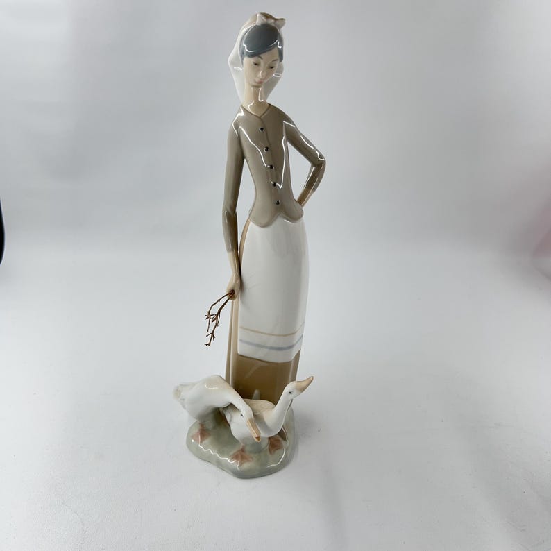 May include: A porcelain figurine of a woman in a long dress and head covering, holding a small branch. Two white geese are at her feet, on a small, green base. The woman's outfit is tan and white, with a gray jacket.