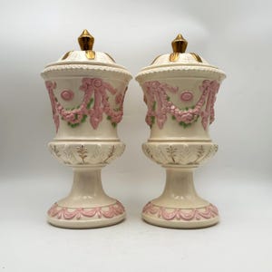 May include: Two matching cream-colored urns with lids, adorned with pink and green floral garlands and gold accents. The urns have a pedestal base and are likely decorative pieces for a home.