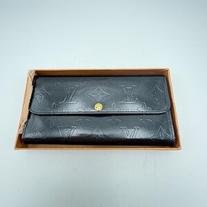 Puede incluir: Cartera de cuero negro con dise&ntilde;o en relieve, cierre de solapa y bot&oacute;n dorado. La cartera se presenta dentro de una caja de cart&oacute;n marr&oacute;n, lo que sugiere un embalaje de regalo o almacenamiento.