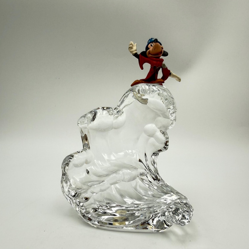 Op de afbeelding: Een heldere glazen sculptuur in de vorm van een golf, bekroond met een klein beeldje van een stripfiguur in een rode jurk en blauwe hoed. Het beeldje zwaait. De sculptuur staat tegen een witte achtergrond.