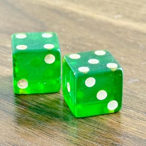 May include: Two transparent green dice with white dots, each showing different numbers. The dice are on a wooden surface, with the light reflecting off the glossy surfaces. A classic game accessory.