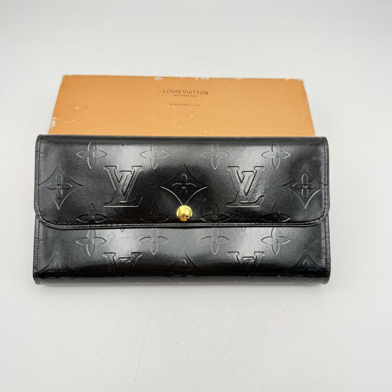 Puede incluir: Cartera de cuero negro con estampado de monograma en relieve y detalles dorados. La cartera tiene un cierre de solapa con un bot&oacute;n dorado. La marca "LOUIS VUITTON" es visible en la caja en el fondo.
