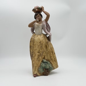 May include: A porcelain figurine of a person with dark hair wearing a white bodice and a long, textured yellow skirt with a green tree detail at the hem. The figure holds a basket of fruit on their head and a small object near their face.