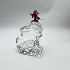 Op de afbeelding: Een heldere glazen sculptuur met een golfachtig ontwerp, bekroond met een klein beeldje van een stripfiguur tovenaar in een rode mantel en blauwe hoed. Het beeldje zwaait met &eacute;&eacute;n hand.