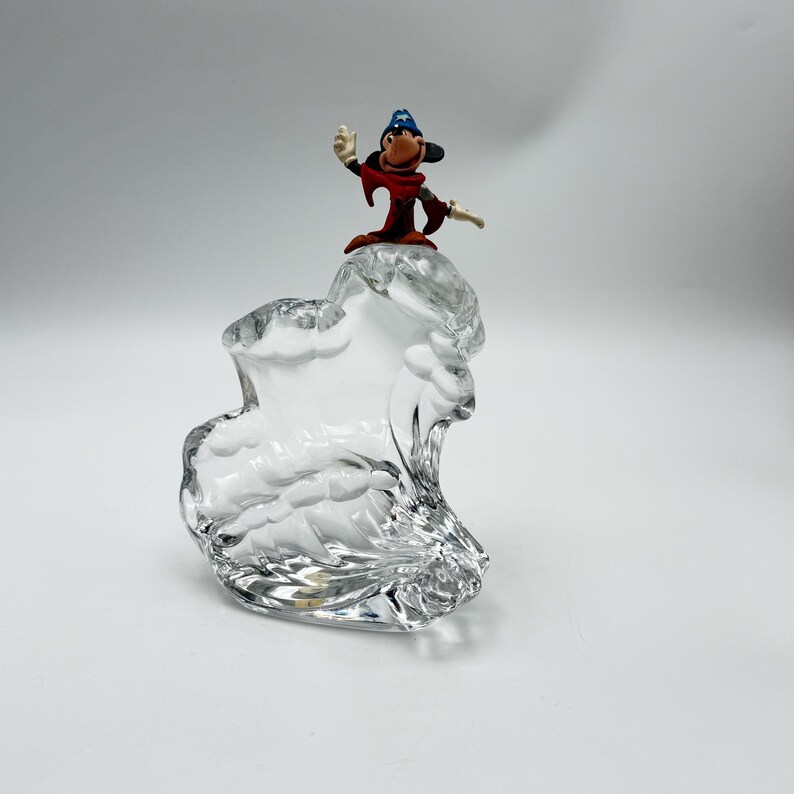 Op de afbeelding: Een heldere glazen sculptuur met een golfachtig ontwerp, bekroond met een klein beeldje van een stripfiguur tovenaar in een rode mantel en blauwe hoed. Het beeldje zwaait met &eacute;&eacute;n hand.