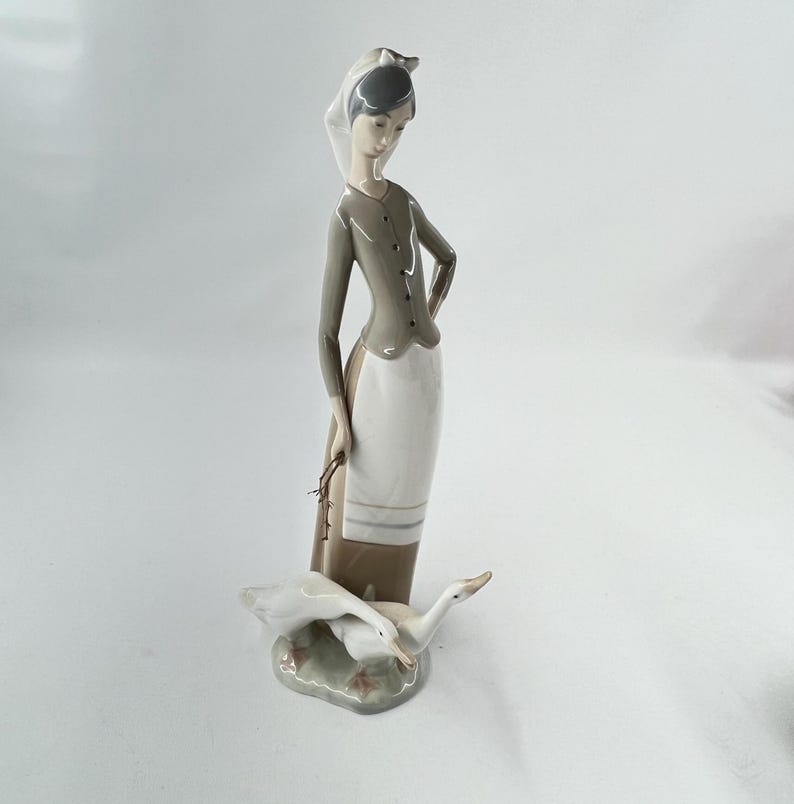 May include: A porcelain figurine of a woman in a long skirt and a headscarf, holding a small bundle. Two white geese are at her feet. The woman's outfit is gray and white. The base is a light green color.