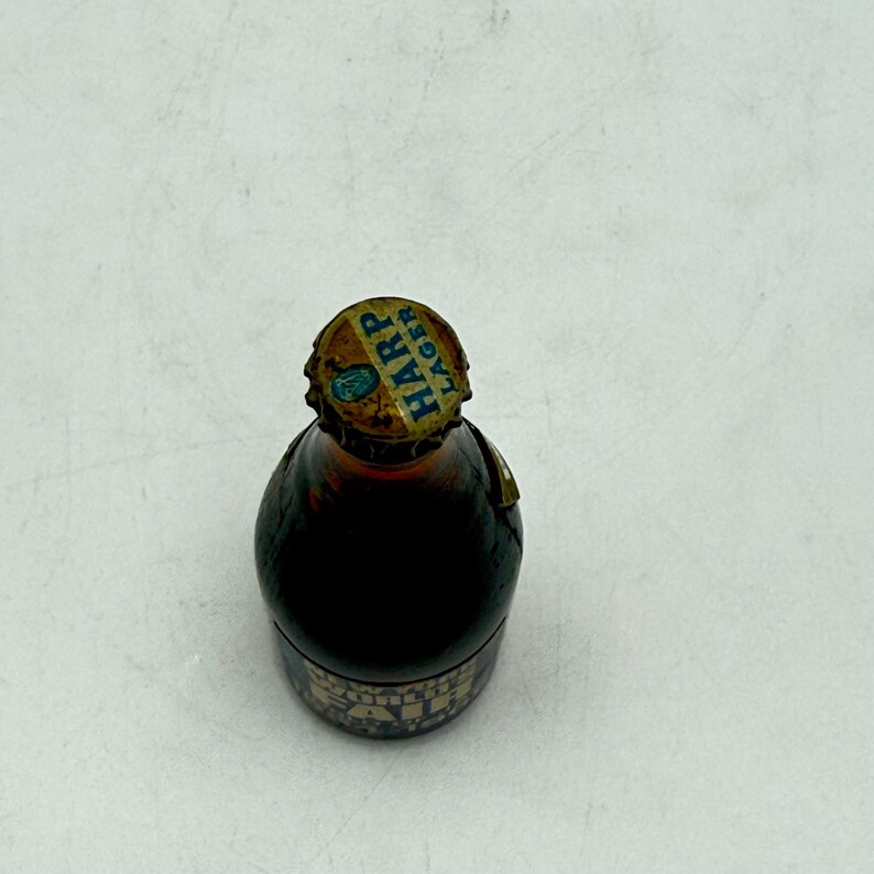 May include: A small, brown glass bottle with a gold and blue Harp Lager bottle cap. The bottle has a dark liquid inside and a decorative label around the base. The bottle is sitting on a white surface.