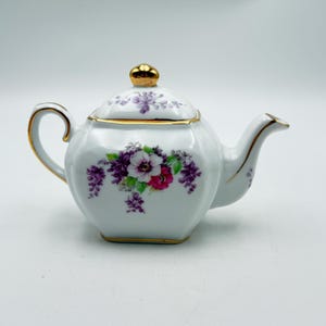 May include: White porcelain teapot with a gold handle, spout, and trim. The teapot features a floral design with purple, pink, and white flowers. The lid is topped with a gold knob.
