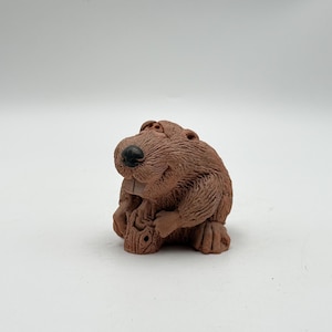 May include: A small, brown, textured clay figurine of a beaver with a black nose and visible front teeth. The beaver is depicted in a seated position, holding a small, carved wooden log between its front paws, showcasing detailed fur markings.