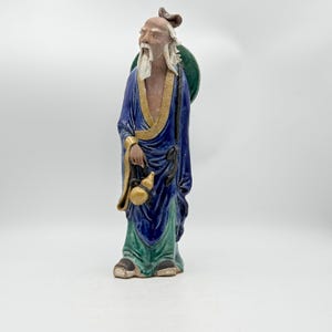 May include: A ceramic figurine of an elderly man in a long blue robe with gold trim and green accents. The figure has a long white beard and carries a gourd and a green disc on his back. The figurine is a decorative item.