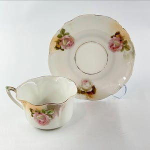 May include: A vintage teacup and saucer set. The set features a scalloped edge design with a cream-colored base. Pink roses with green leaves are painted on both the cup and saucer, with gold trim accents.