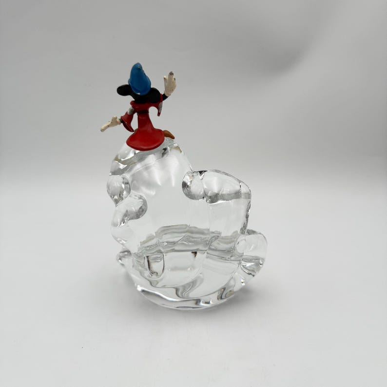 Op de afbeelding: Een heldere glazen asbak met een cartoonfiguur. Het beeldje draagt een rode jurk en een blauwe hoed. De asbak heeft een unieke vorm. Een decoratief en functioneel item voor in huis.