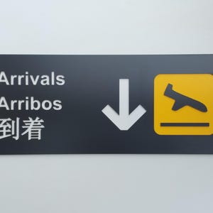May include: A black sign with white text that reads "Arrivals, Arribos, 到着" and a white arrow pointing down. A yellow square with a black airplane silhouette is on the right side of the sign.