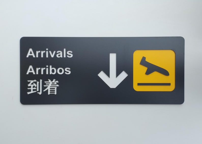May include: A black sign with white text that reads "Arrivals, Arribos, 到着" and a white arrow pointing down. A yellow square with a black airplane silhouette is on the right side of the sign.