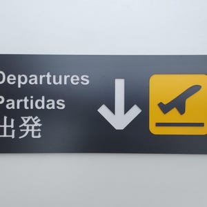 May include: Black sign with white text that reads "Departures, Partidas, 出発" with a white downward arrow and a yellow square with a black airplane taking off.