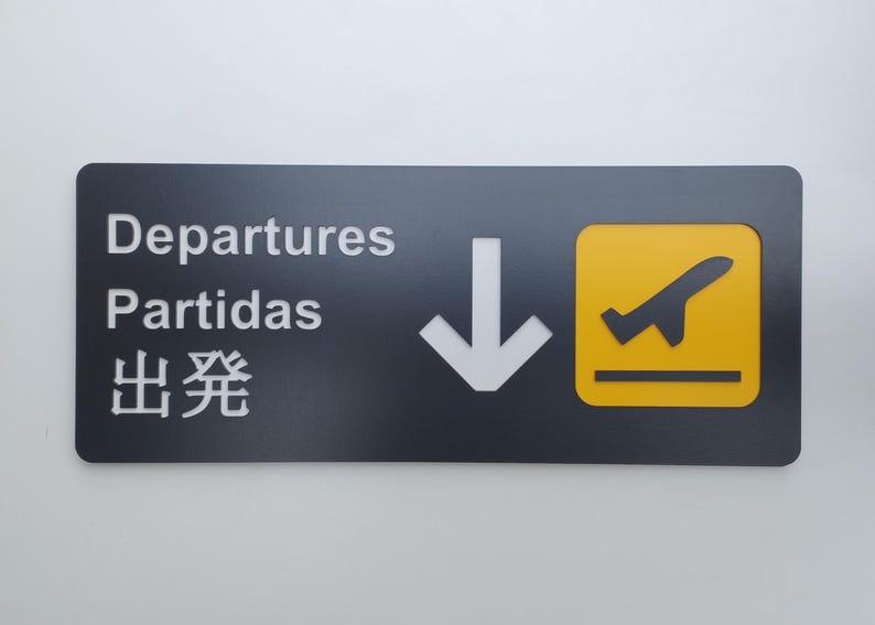 May include: Black sign with white text that reads "Departures, Partidas, 出発" with a white downward arrow and a yellow square with a black airplane taking off.