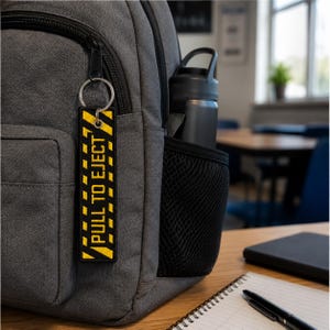 May include: A rectangular fabric keychain featuring a black and yellow hazard stripe pattern with the text PULL TO EJECT printed in bold yellow letters. The keychain is attached to a silver metal split ring and a metal grommet, hanging from a gray backpack.
