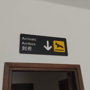 May include: Black and yellow sign with white text and a white arrow pointing down. The sign reads "Arrivals, Arribos, 到達".