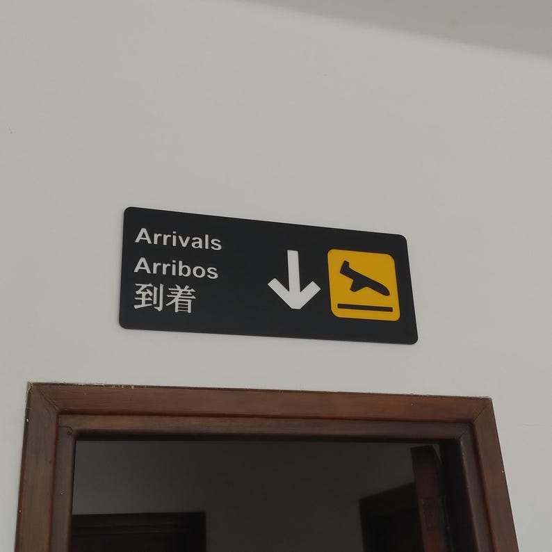 May include: Black and yellow sign with white text and a white arrow pointing down. The sign reads "Arrivals, Arribos, 到達".