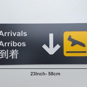 May include: Black and yellow airport sign with white text that reads "Arrivals", "Arribos", and "到着".  The sign also has a white downward arrow and a yellow airplane icon. The sign is 23 inches wide and 9.4 inches tall.