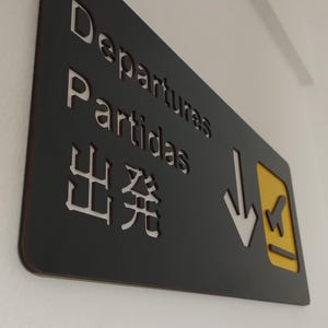 May include: Black sign with white text that reads "Departures, Partidas, 出発" with a yellow arrow pointing down and a small airplane icon.
