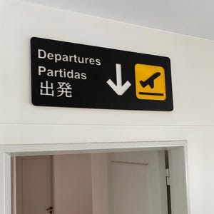 May include: Black and yellow sign with white text that reads "Departures, Partidas, 出発" with a white arrow pointing down and a yellow airplane icon.
