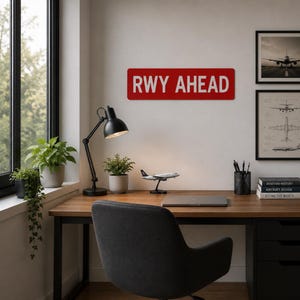 Puede incluir: Un letrero rectangular rojo con texto blanco que dice RWY AHEAD. El letrero está montado en una pared sobre un escritorio de madera que cuenta con una lámpara de escritorio negra, una pequeña planta en maceta, un modelo de avión, una computadora portátil y un portalápices.