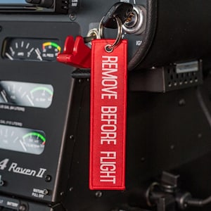 Könnte beinhalten: Ein rotes, rechteckiges Schlüsselanhänger-Etikett mit den Worten "REMOVE BEFORE FLIGHT" in Weiß gestickt. Das Etikett ist an einem Schlüssel und einem roten Sicherheitsclip befestigt, vor einer dunklen Instrumententafel.