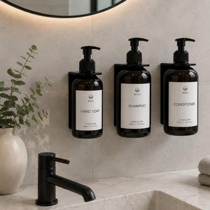 Könnte beinhalten: Drei wandmontierte Braunglasflaschen mit schwarzen Pumpdosierern, beschriftet mit Hand Soap, Shampoo und Conditioner. Jede Flasche hat ein weißes Etikett mit dem Text MIZU, Home & Spa und 500ml/16.9fl.oz. Die Flaschen werden von schwarzen Halterungen gehalten.