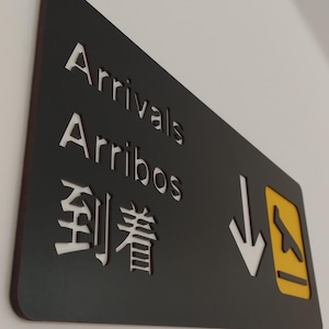 May include: Black sign with white text that reads "Arrivals Arribos 到着" with a yellow and white airplane icon and a white arrow pointing down.