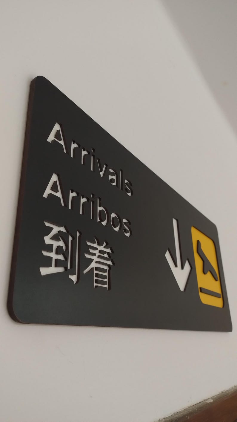 May include: Black sign with white text that reads "Arrivals Arribos 到着" with a yellow and white airplane icon and a white arrow pointing down.