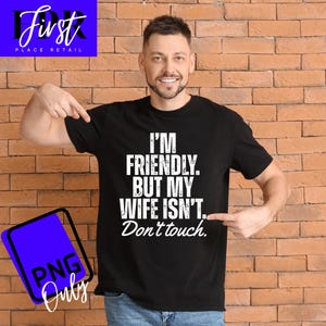 Puede incluir: Camiseta negra con el texto blanco "I'M FRIENDLY. BUT MY WIFE ISN'T. Don't touch.". Una persona señala el texto. El fondo es una pared de ladrillos.